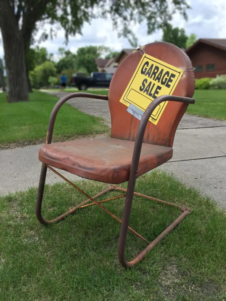 A metal chair sits on a lawn with a "Garage Sale" sign on it