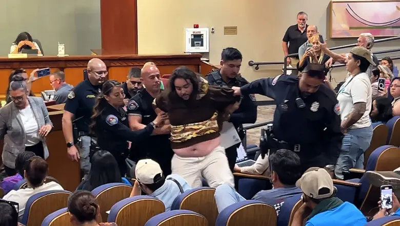 A man is forcefully removed by police following an explosive public comment period at City Hall. Behind the commotion, Councilor Rogers is seen covering her face, while Councilor Telles follows the group out to speak with the man and defuse the confrontation. (Jesse Jones)