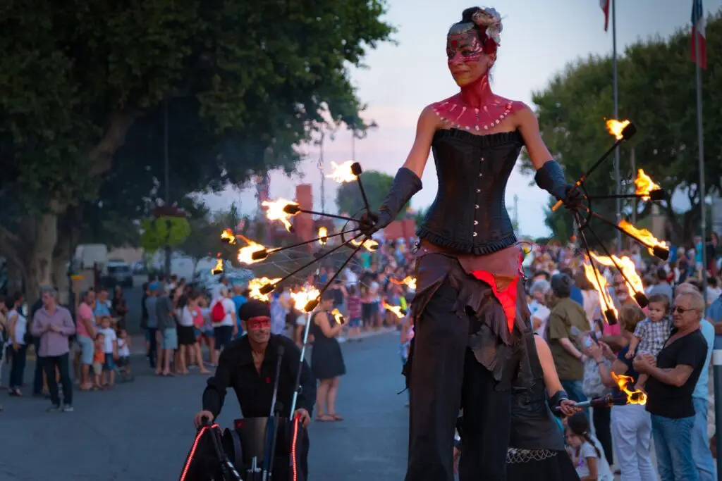 A woman in a corset walks down a street carrying fire fans.