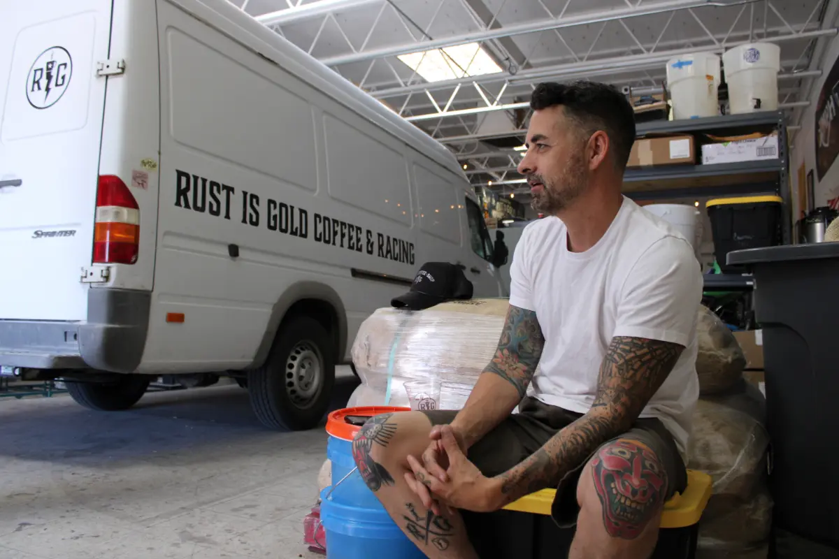 A man sits in front of a white van marked Rust is Gold Coffee & Racing