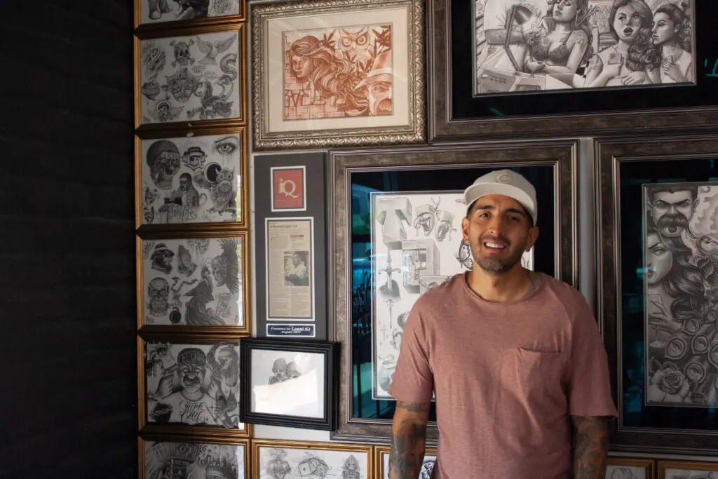 A man in a baseball cap stands in front of a wall filled with tattoo flash art.