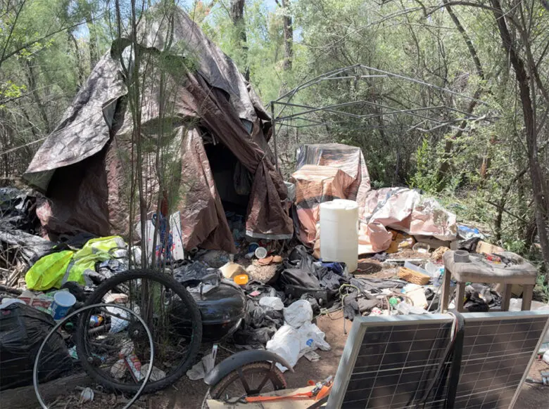 A makeshift tarp tent stands among debris, bicycles and a solar panel at a Bosque encampment before crews cleared the site. (Courtesy Bernalillo County)
