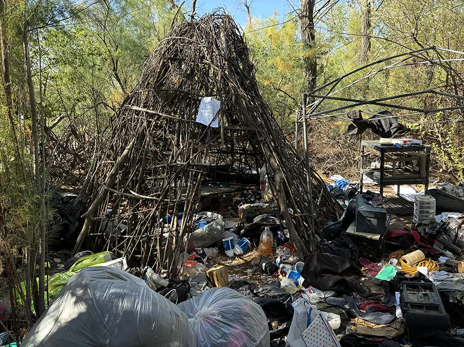 A shelter constructed from tree branches is surrounded by trash bags, plastic bins, household items and a canopy frame in the Bosque. (Courtesy Bernalillo County)