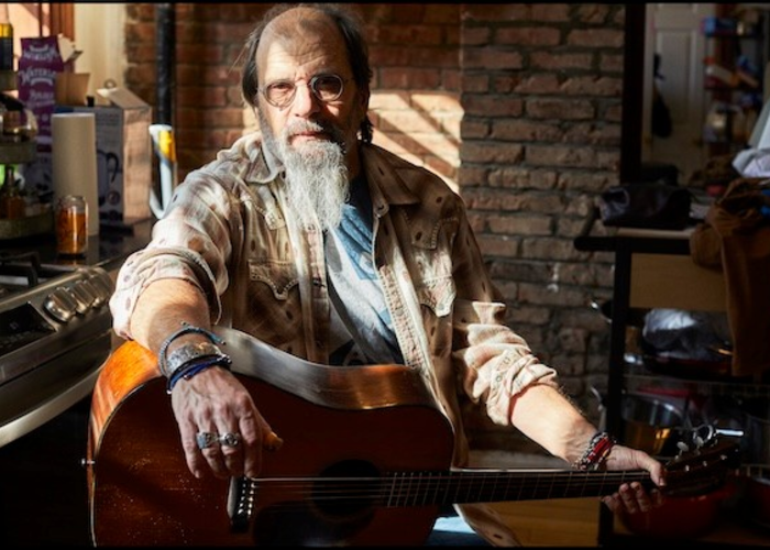 Bearded musician Steve Earle sits in a chair in the sunlight.