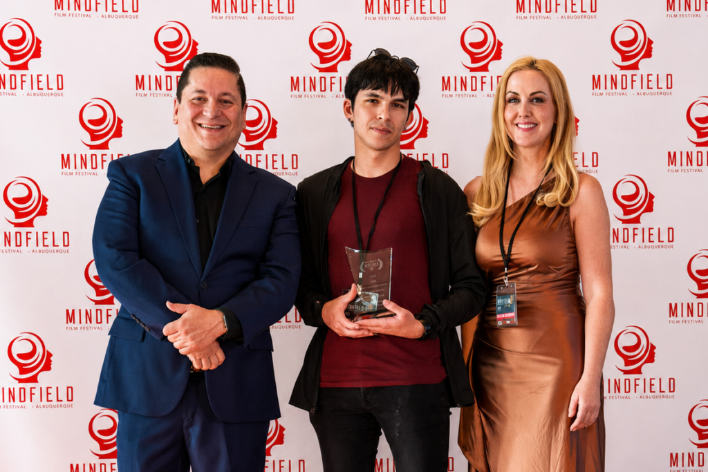 Two well dressed men and a woman in a gold dress hold an award while posing in front of a photo backdrop.