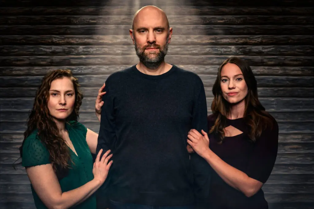 A tall man in a dark shirt stands against a brick wall between two women.
