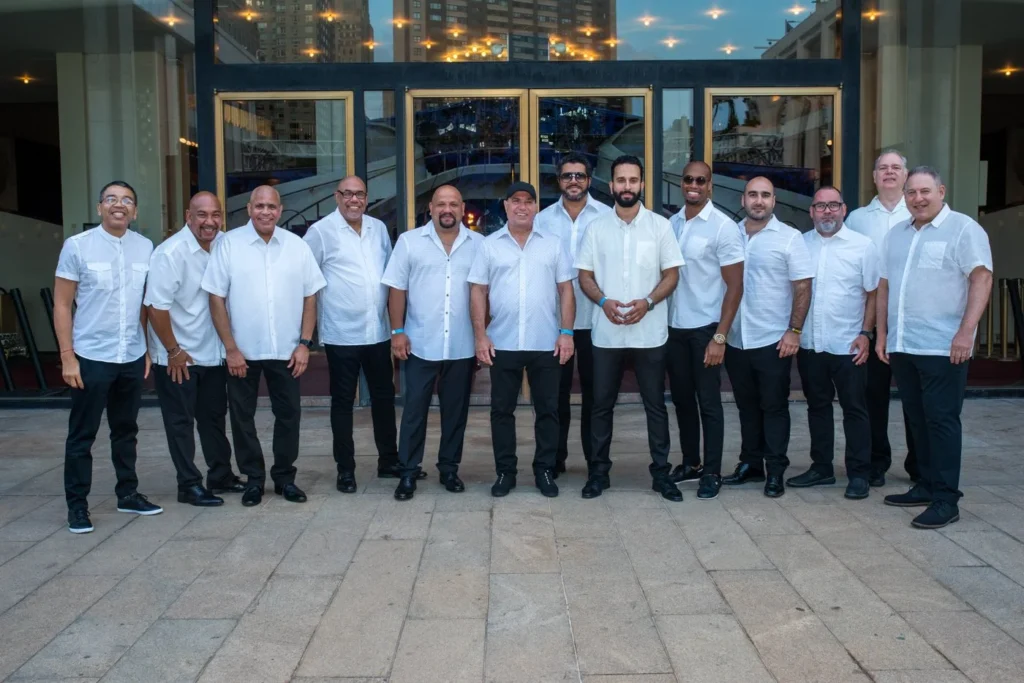 A large group of musicians in white Guanabara shirts stands in front of a music venue.