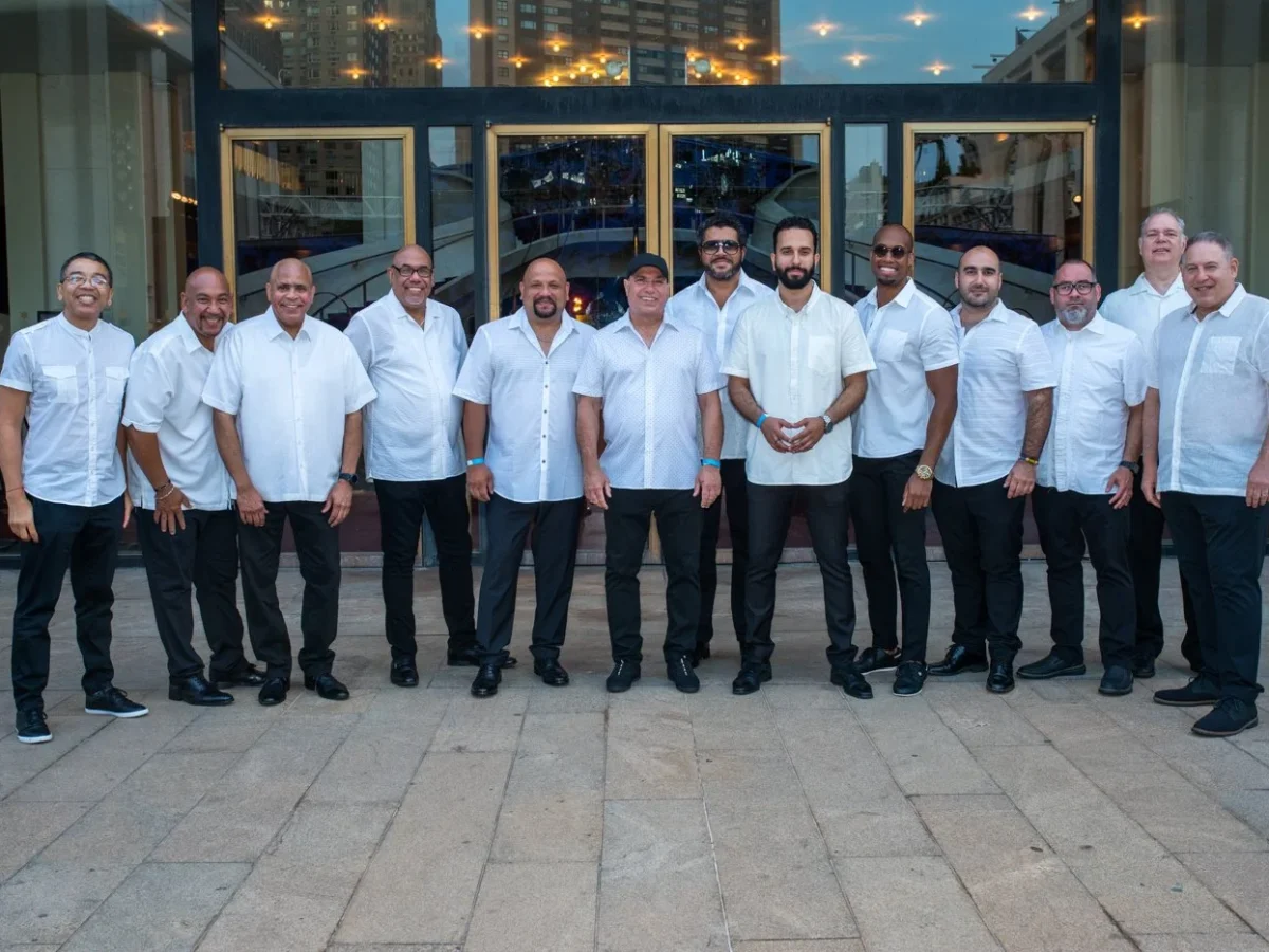 A large group of musicians in white Guanabara shirts stands in front of a music venue.